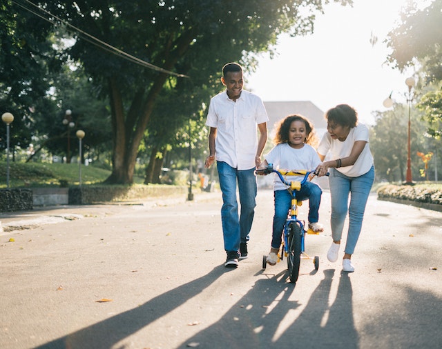 two parents helping their child learn how to ride their bike on a sunny street